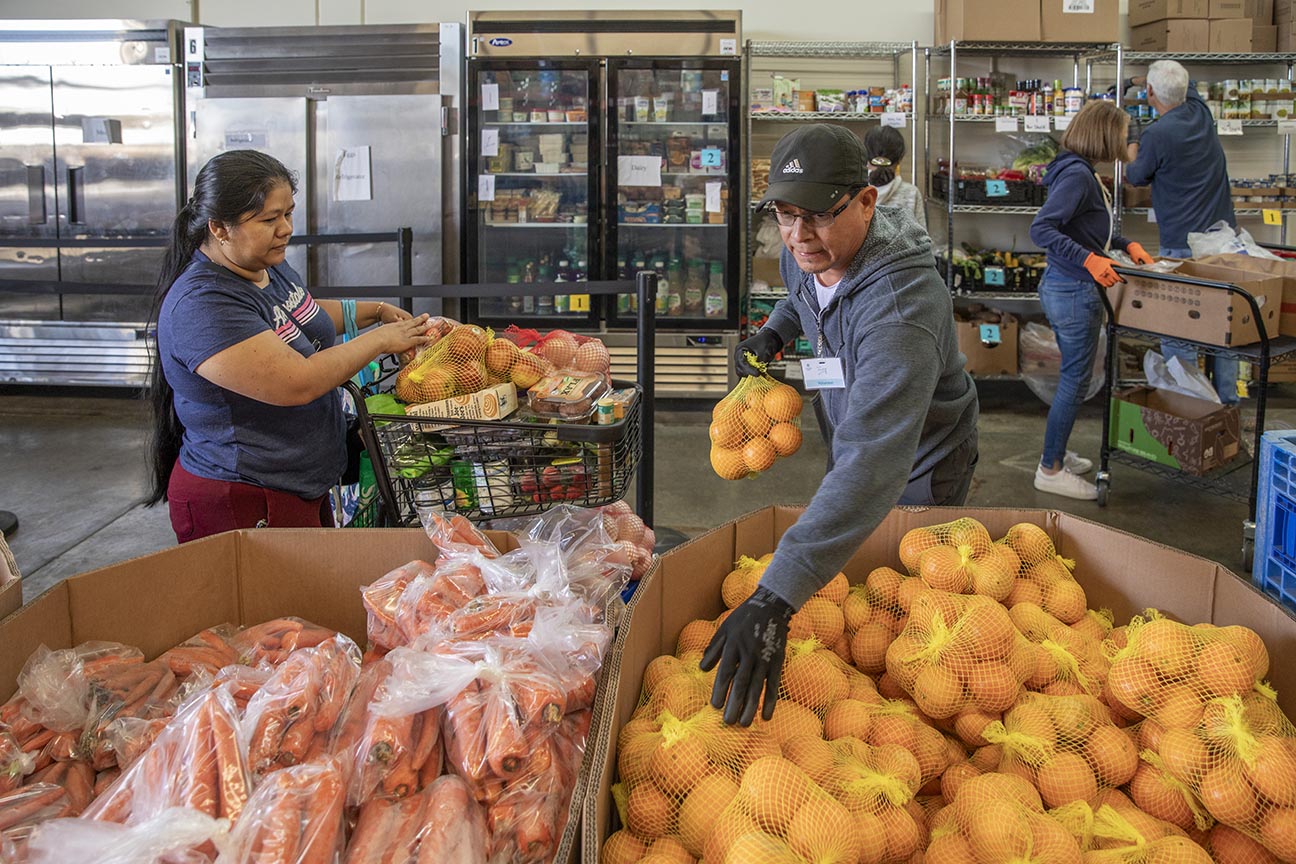 Man sorting surplus bags of fruit next to woman with shopping cart full of food